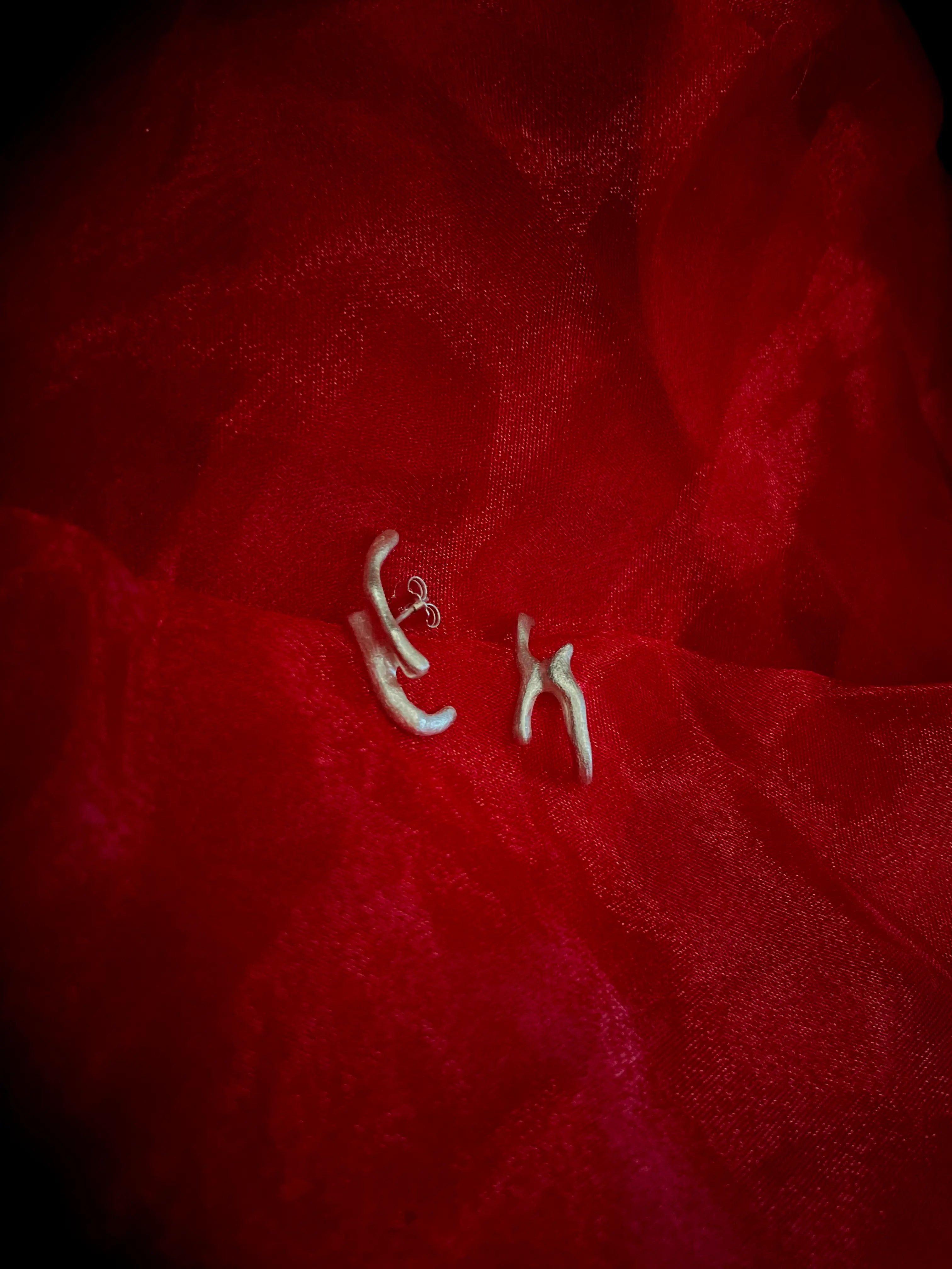 Détail macro de la boucle d'oreille Arki en argent 925, posée sur un tissu rouge texturé. Bijou artisanal Synonys.